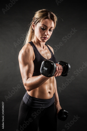 Fitness in gym, sport and healthy lifestyle concept. Beautiful athletic woman showing her trained body on black background. Bodybuilder female model training biceps muscles with dumbbell.