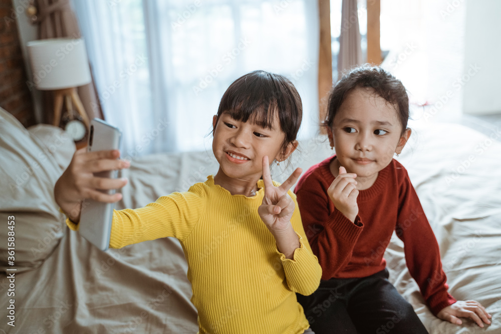 two little asian girls smile while taking a selfie together with a smartphone camera when sitting on the bed two little asian girls smile while taking a selfie together with a smartphone camera when sitting on the bed