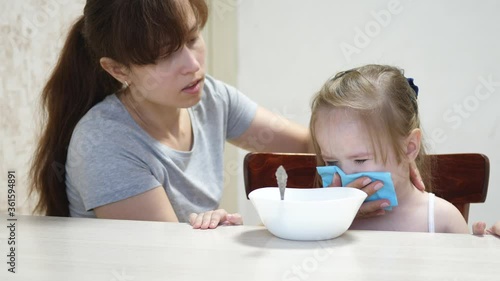 Mom calms a capricious child sitting at the table. Parent wipes kid drool and snot with a napkin. Teamwork. Baby doesn t want to eat porridge photo