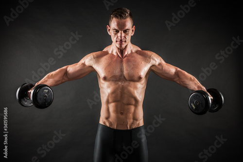 Fitness in gym, sport and healthy lifestyle concept. Handsome athletic man showing his trained body on black background. Bodybuilder male model training biceps muscles with dumbbell. © Maksim Toome