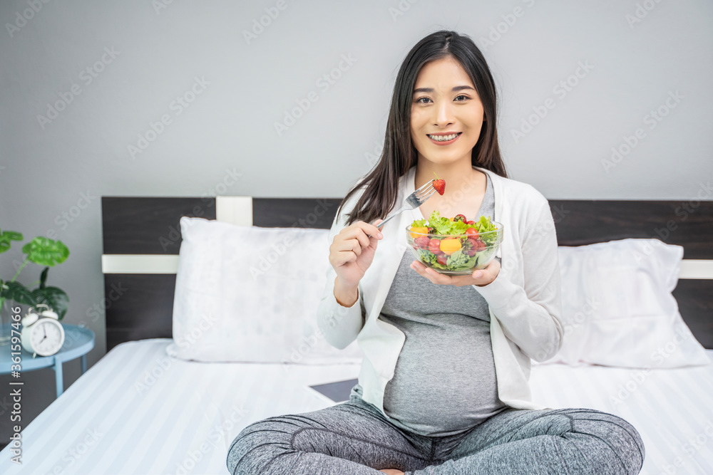 Beautiful asian pregnant woman holding a bowl of salad of vegetables and fruits, using a fork eating a strawberry. Eating healthy having lunch sitting on the white bed, relaxing and resting in bedroom Beautiful asian pregnant woman holding a bowl of salad of vegetables and fruits, using a fork eating a strawberry. Eating healthy having lunch sitting on the white bed, relaxing and resting in bedroom