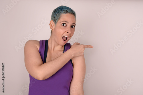 Cheerful surprised woman showing product. Stylish bright hipster girl with blue short hair in violet top points to the side isolated on light background. Copy space for text or advertising