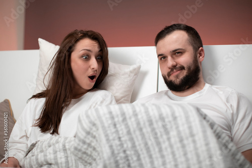 Beautiful loving married couple sitting in bed. Boyfriend raised blanket showing what under it for excited surprised girlfriend. Self-confident man in bedroom, happy sex life, sexual activity concept
