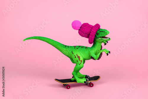 cute green toy dinosaur in knitted hat driving skateboard on a pink background, funny greeting card © dvulikaia