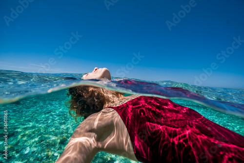 Carefree and relaxed, close up, young athletic woman effortlessly floats on her back in tropical ocean waters under blue sky wearing red one piece bathing suit
