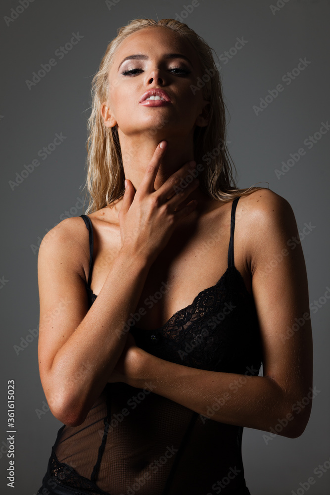 Naklejka premium Portrait of blond young woman with beautiful makeup and hairstyle in a black lace bodysuit stands on gray background. Fashion model poses in elegant clothes in studio