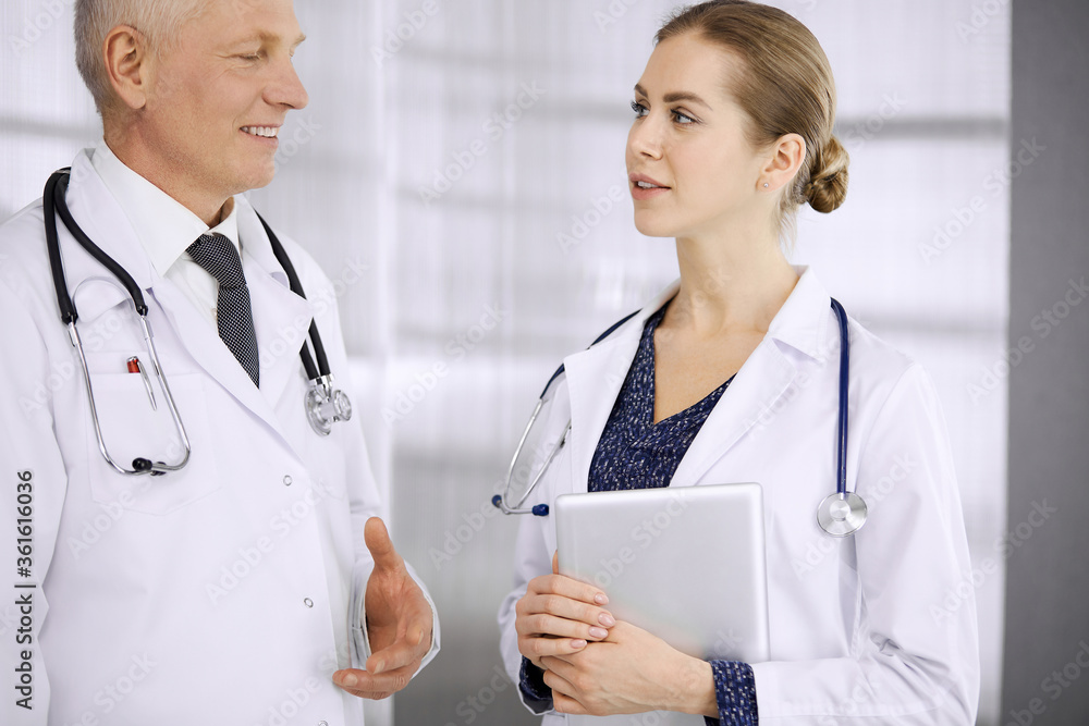 Fototapeta premium Elderly confident doctor and woman physician discussing current disease therapy while standing in clinic. Teamwork in hospital. Medicine and healthcare concept