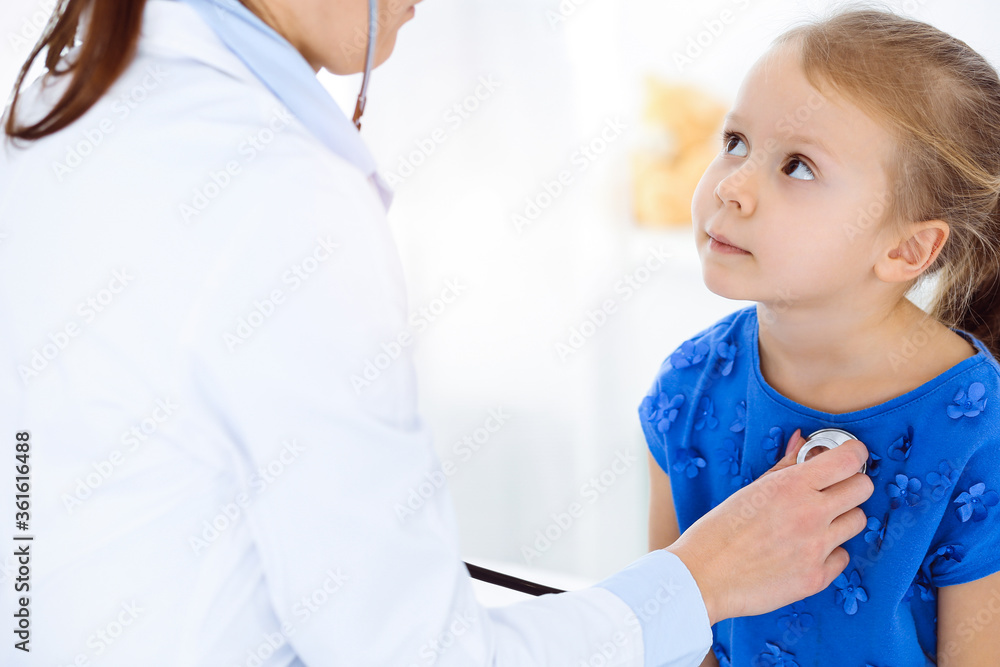 Doctor examining a child by stethoscope in sunny clinic. Happy smiling girl patient dressed in blue dress is at usual medical inspection Doctor examining a child by stethoscope in sunny clinic. Happy smiling girl patient dressed in blue dress is at usual medical inspection