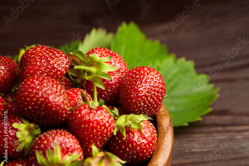 Strawberries with green leaves in wooden bowl on brown wooden table. Red ripe berries