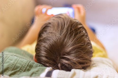 Adorable boy sitting on the sofa in the living room and playing with smartphone. Child learning how to use smartphone. Boy texting on the phone. - technology and lifestyle concept.