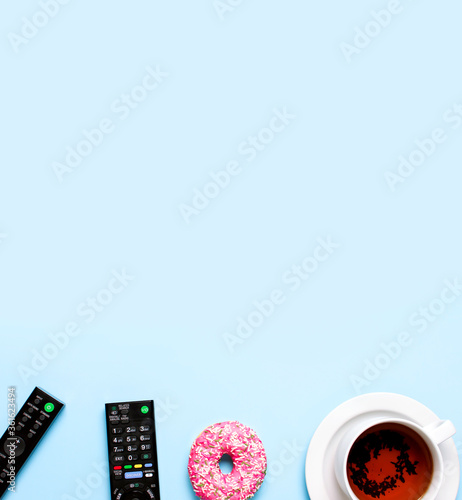 Black TV  audio remote control  cup of tea  pink sweet donut on blue background flat lay top view copy space. Minimalistic background with remote control. Concept of spending time in front of the TV