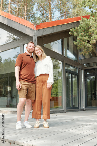 Positive dreamy young couple standing against new house with glass walls and embracing while thinking of future