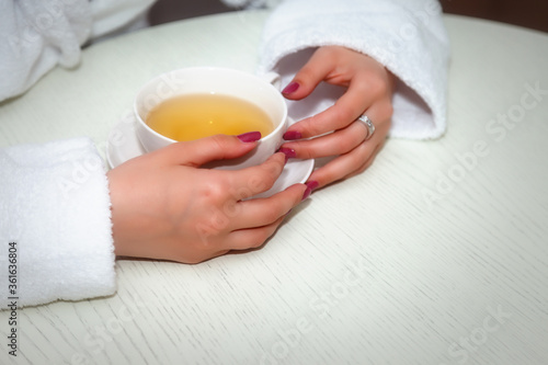 Cup of tea in womans hands. Girl in white coat at table. Young charming female is sitting in hotel room and holding Cup of tea. Tea ceremony concept. Space for an inscription or logo