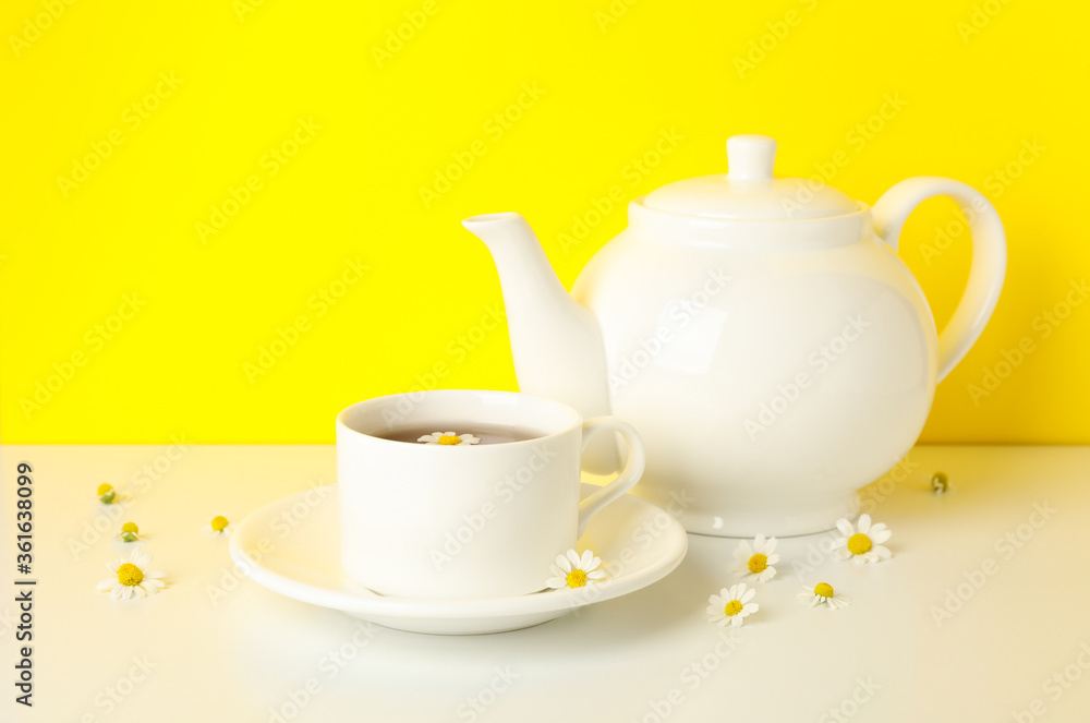 Teapot, cup of tea and chamomile on white table against yellow background Teapot, cup of tea and chamomile on white table against yellow background