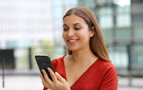 Portrait of beautiful casual business woman sending message with mobile phone chatting app with modern skyscrapers on the background