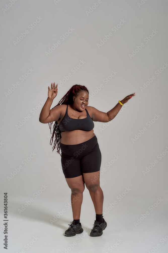 Foto de I love my curves. Full length shot of plump, plus size african  american woman in sportswear having fun, dancing in studio over grey  background do Stock
