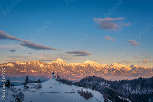 Gorgeous and colorful sunrise over Jamnik church with Alps mountains range in the distance. Famous landmark in Slovenia in wintertime. Landscape covered with snow