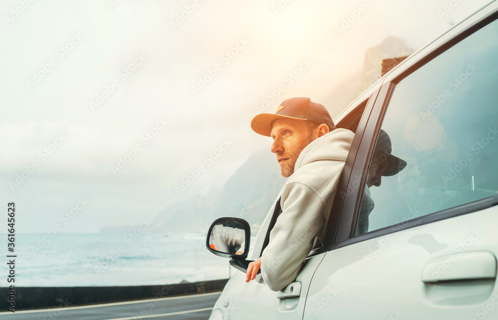 Naklejka premium Man dressed warm hoodie and baseball cap driving his new car on the lonely road leading by the sea coast. Happy auto owner concept image.
