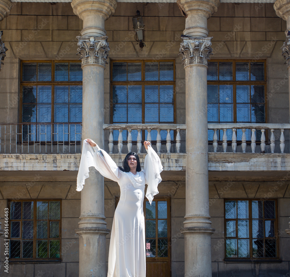 Drag queen posing close to old building in white dress. A wig with Black long hair and make up. High quality photo