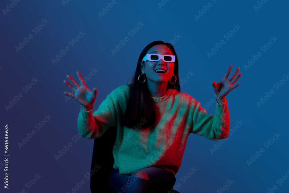 Excited woman wearing cinema 3d glasses in blue and red light is sitting on a chair
