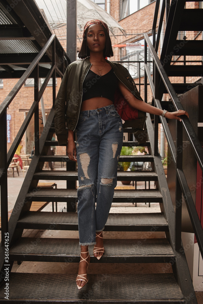 Full length trendy fashion portrait of young beautiful stunning african woman in jeans and black top walking staircase. Fashion stylish model.