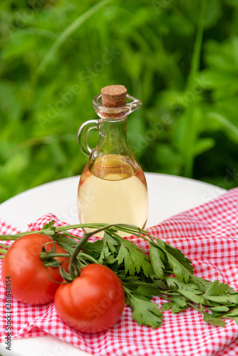 Olive oil in a glass bottle with tomatoes on a branch and parsley in the fresh air on a background of green grass.