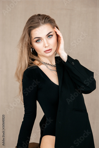 Portrait of a beautiful girl in a black jacket standing sideways, leaning on one hand, the second hand at the face. Metal accessories.Black clothes.Beige background.