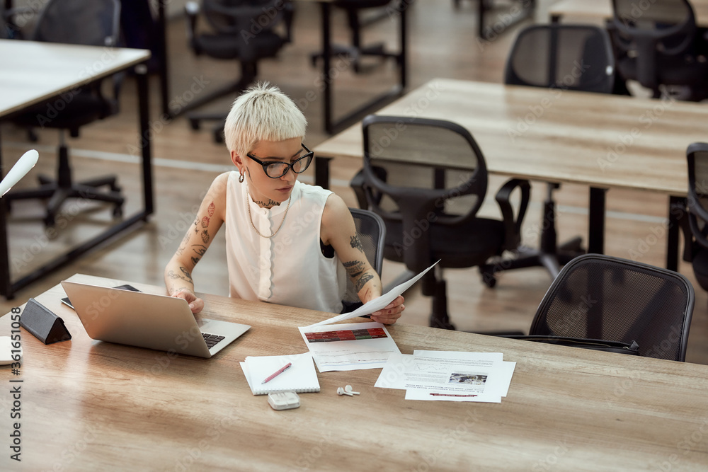 Business analysis expert. Young and cute business woman with short haircut analyzing documents and working with a laptop while sitting at her desk in the office Business analysis expert. Young and cute business woman with short haircut analyzing documents and working with a laptop while sitting at her desk in the office