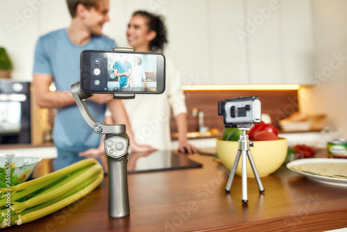 Happy couple, vegetarians recording a video on camera and smartphone while going to prepare a healthy meal in the kitchen together. Vegetarianism, healthy food, blog concept