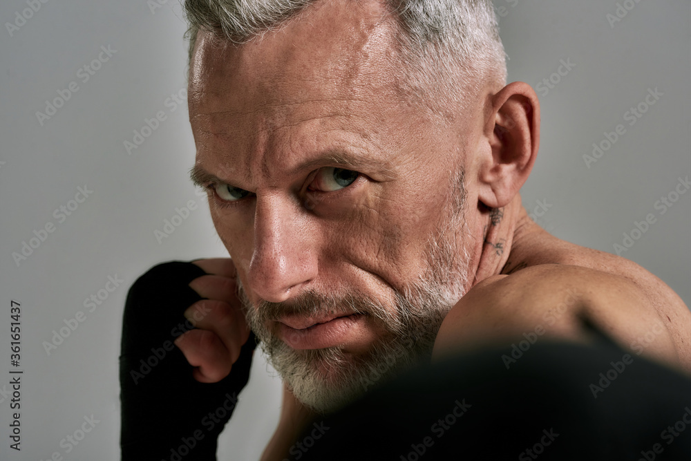 Close up of middle aged athletic man, kickboxer looking agressive while boxing, training in studio over grey background. Muay Thai, Boxing or Kickboxing concept Close up of middle aged athletic man, kickboxer looking agressive while boxing, training in studio over grey background. Muay Thai, Boxing or Kickboxing concept