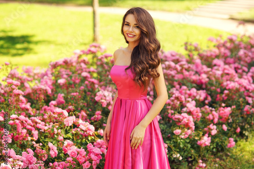 Princess. Young beautiful pretty woman posing in long evening luxury dress against bushes with pink roses on a sunny summer day. Vogue style fashion sensual portrait