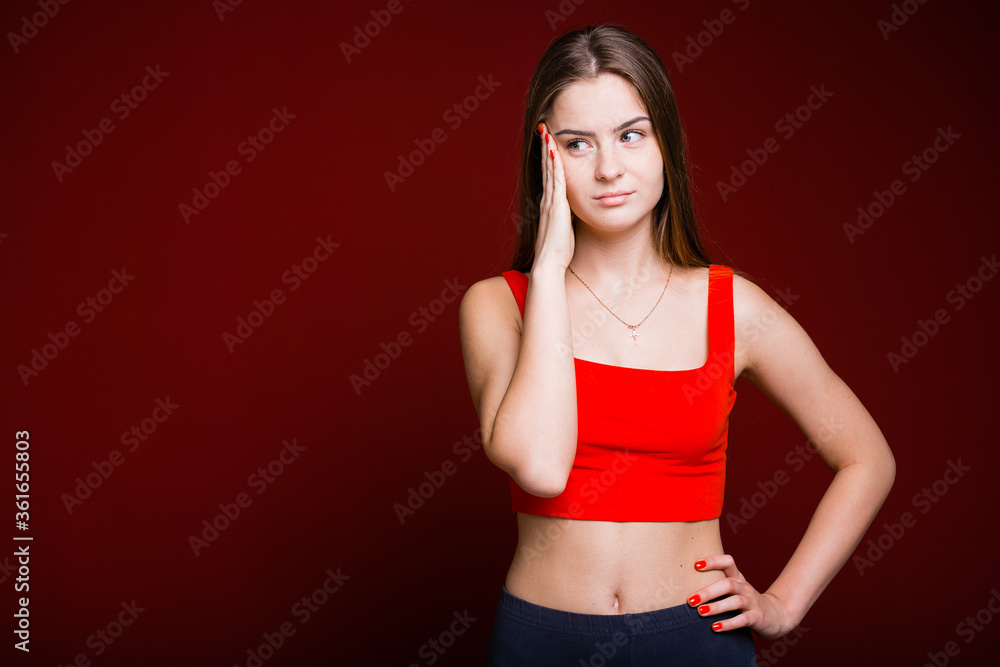 American girl on a red background holds her hand near the temple because of a headache and looks to the side while holding her hand on the waist