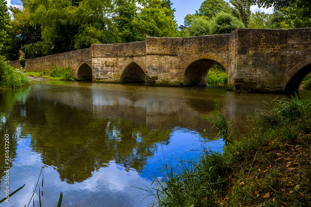A panorama view down the River Ise towards the bridge and ford in the town of Geddington, UK in summer