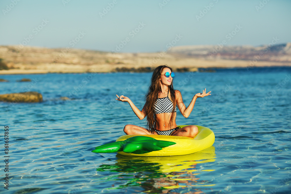 Summer lifestyle portrait of pretty happy young girl with tanned sexy body. Doing yoga, smiling and sitting on air mattress in the sea at the tropical island beach. Wearing stylish bikini, sunglasses Summer lifestyle portrait of pretty happy young girl with tanned sexy body. Doing yoga, smiling and sitting on air mattress in the sea at the tropical island beach. Wearing stylish bikini, sunglasses