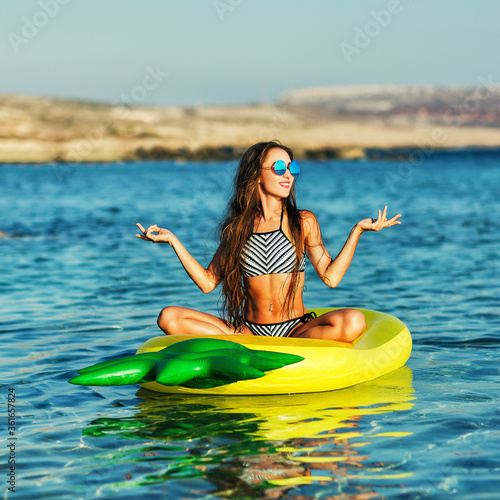 Summer lifestyle portrait of pretty happy young girl with tanned sexy body. Doing yoga, smiling and sitting on air mattress in the sea at the tropical island beach. Wearing stylish bikini, sunglasses