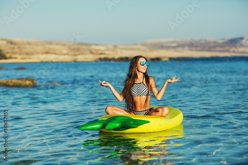 Summer lifestyle portrait of pretty happy young girl with tanned sexy body. Doing yoga, smiling and sitting on air mattress in the sea at the tropical island beach. Wearing stylish bikini, sunglasses © Olesya Kuprina