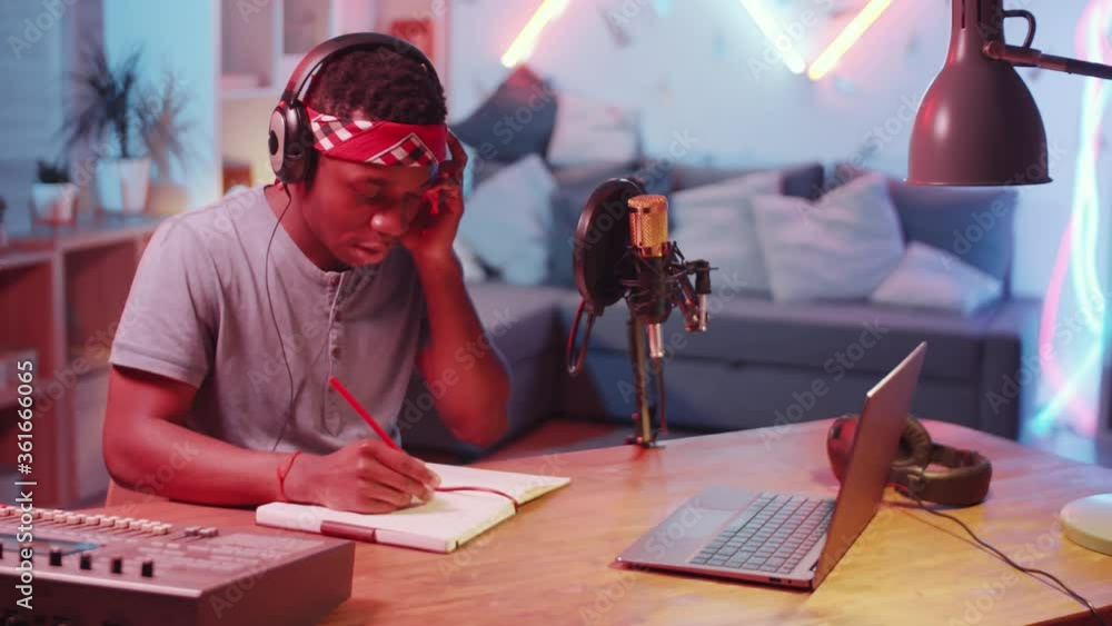 Young African American man in headphones sitting at desk in home recording studio, moving his head to music and writing lyrics in notepad while working on new song