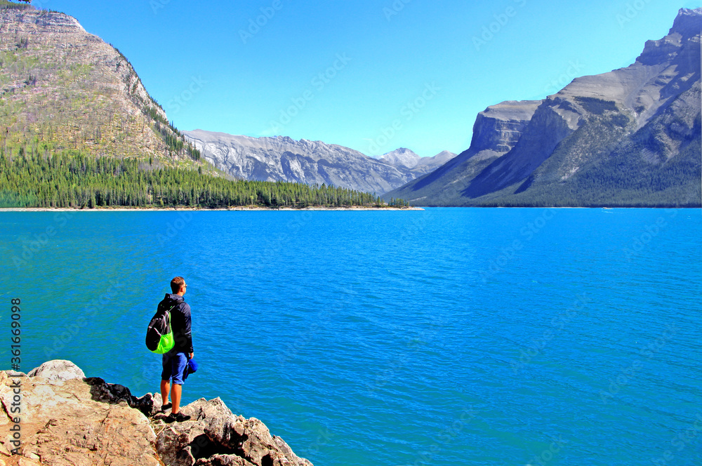 Obraz premium Man with backpack standing on the rocky coast of Minnewanka lake on beautiful sunny day in Banff National Park, AB.