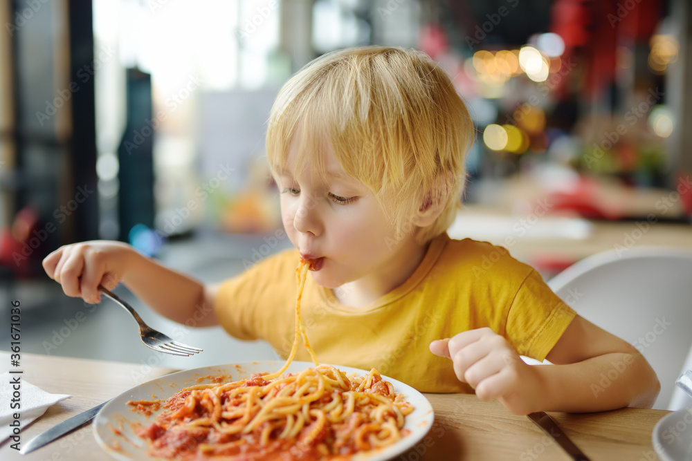 Preschooler boy eating pasta with tomatoes in cafe or restaurant. Healthy/unhealthy food for little kids Preschooler boy eating pasta with tomatoes in cafe or restaurant. Healthy/unhealthy food for little kids