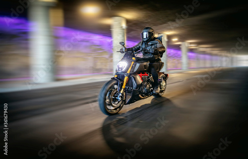 Motorcyclist in full black leathers and helmet riding fast through tunnel with columns and purple light in background