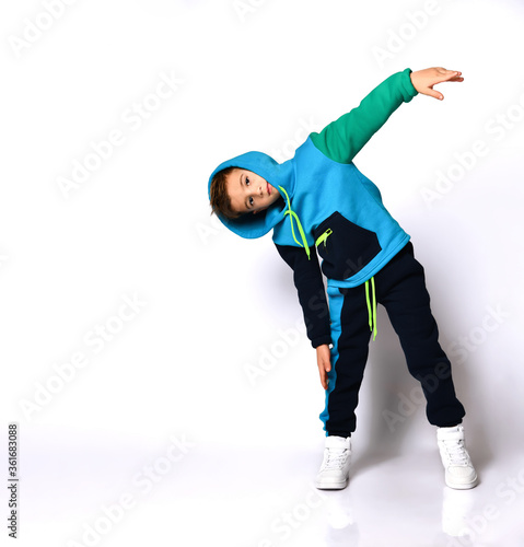 Little brunet boy in colorful sport suit and sneakers. He has put his hood on, performing exercises, posing isolated on white