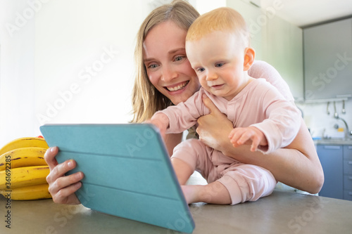 Happy joyful mom and baby daughter talking to family in kitchen, using tablet for video call, smiling at screen together. Child care or online communication concept