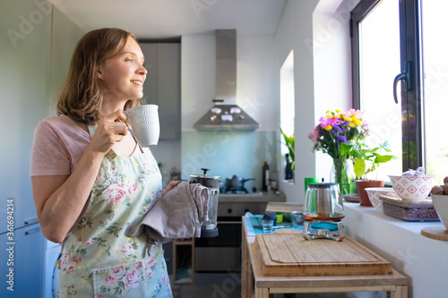 Happy dreamy housewife wearing apron  drinking tea and looking out window in her kitchen. Cooking at home and tea break concept