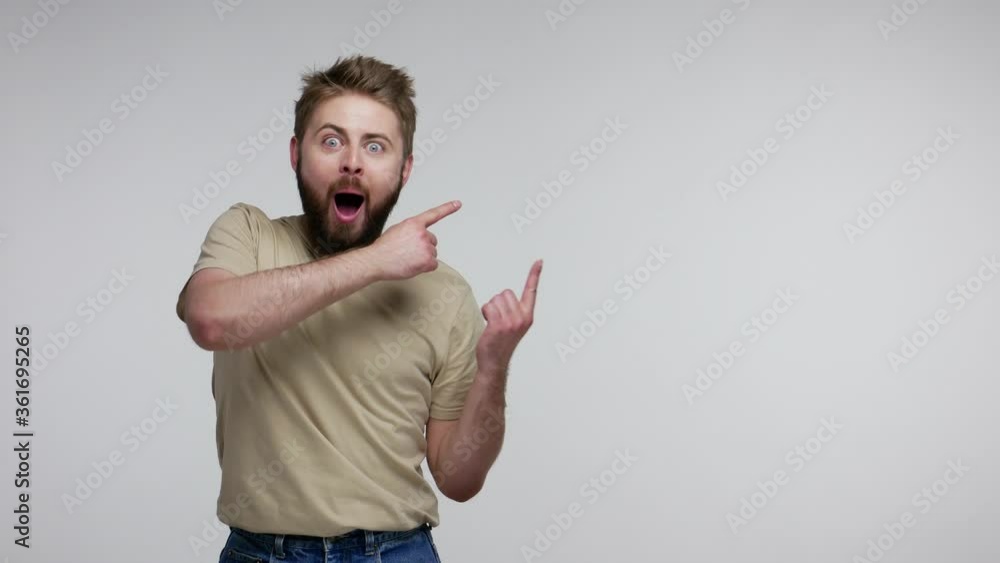 Surprised bearded guy in t-shirt pointing aside and looking with open mouth amazed shocked extremely excited, showing blank copy space for unbelievable advertise. studio shot isolated, gray background Surprised bearded guy in t-shirt pointing aside and looking with open mouth amazed shocked extremely excited, showing blank copy space for unbelievable advertise. studio shot isolated, gray background
