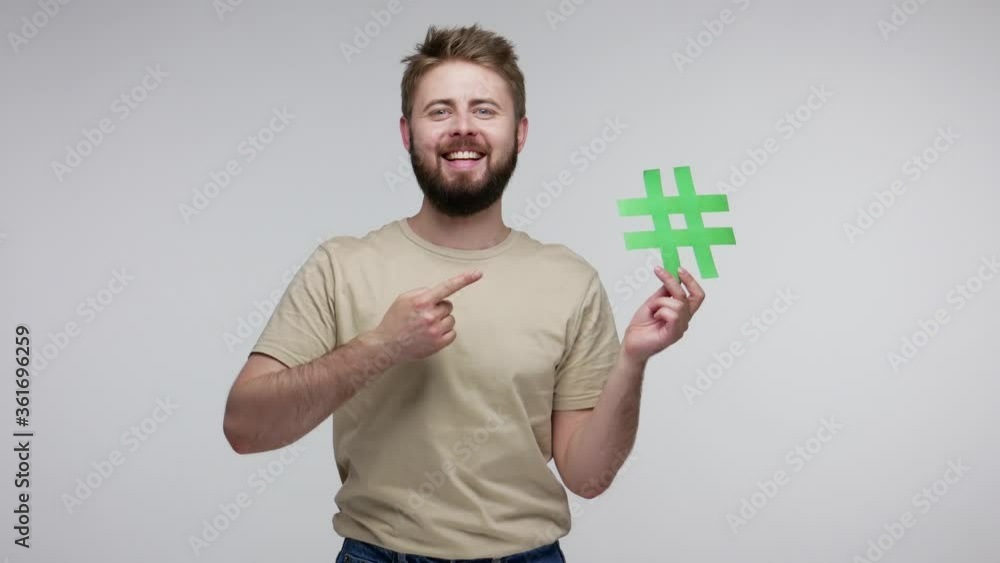 Read tagged message! Cheerful bearded guy pointing hashtag symbol, smiling at camera, showing hash with copy space for viral internet post, popular idea. indoor studio shot isolated on gray background