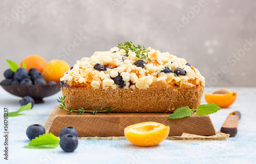 Blueberry and apricot crumble cake decorated with thyme on wooden cutting board. Blue stone background.