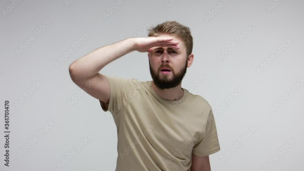 Attentive bearded man carefully looking far away inspecting horizon, holding hand over eyes peering distance, pointing happily to camera when discovered you. studio shot isolated on gray background Attentive bearded man carefully looking far away inspecting horizon, holding hand over eyes peering distance, pointing happily to camera when discovered you. studio shot isolated on gray background