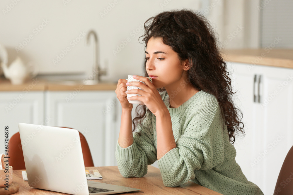 Beautiful young woman drinking hot tea while working on laptop at home Beautiful young woman drinking hot tea while working on laptop at home