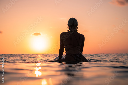 Portrait from the water of surfer girl with beautiful body on surfboard in the ocean at colourful sunset time