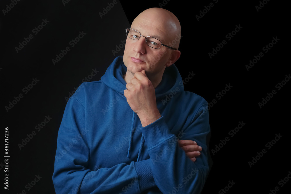 Portrait of bald and clean shaven man with glasses dressed in blue hoodie on a black background looking at viewer. Hand at his face. Portrait of bald and clean shaven man with glasses dressed in blue hoodie on a black background looking at viewer. Hand at his face.